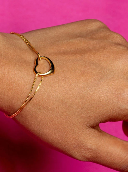 Gold bracelet with a heart-shaped clasp on a wrist against a pink background